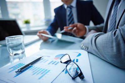 People looking at papers, sitting at a desk with glasses on the desk, pen, charts and glass of water